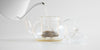 Side view of water being added to a glass teapot, with leaves in the nested glass infuser.