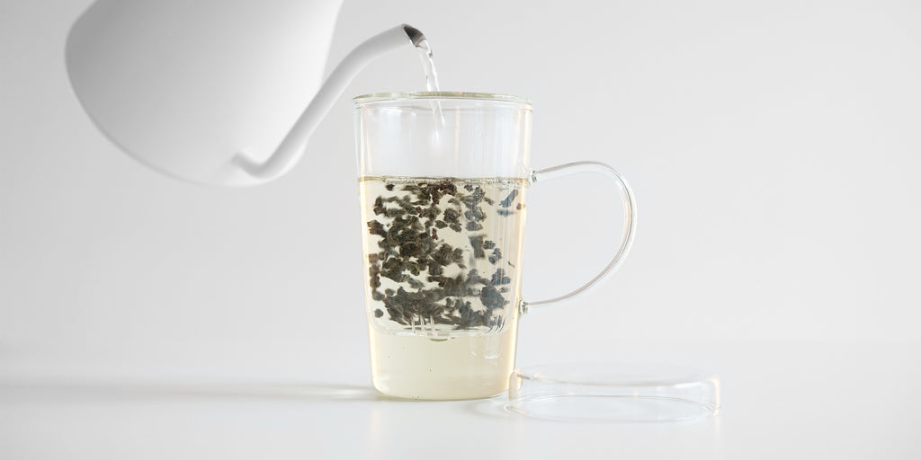 Side view of a glass up with a tea infuser. Water is being added, causing the leaves to churn. A glass lid is placed off to the side.