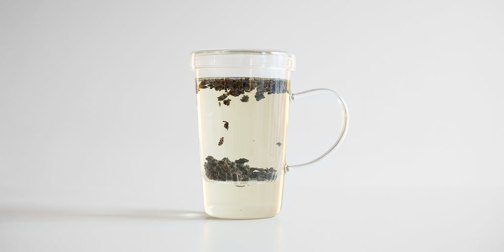 Side view of a glass cup with tea brewing in it. Tea leaves are rising and falling in the inset glass infuser.