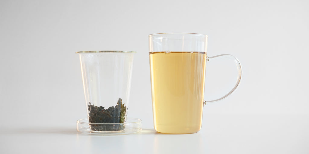Side view of a glass cup with tea in it, a glass infuser on the side filled with tea leaves resting on a glass lid.