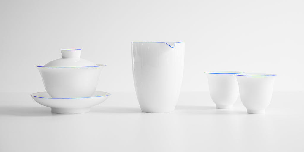A set of matching white porcelain teaware with blue rings around the lips. From left to right, a gaiwan, a server, and two teacups.