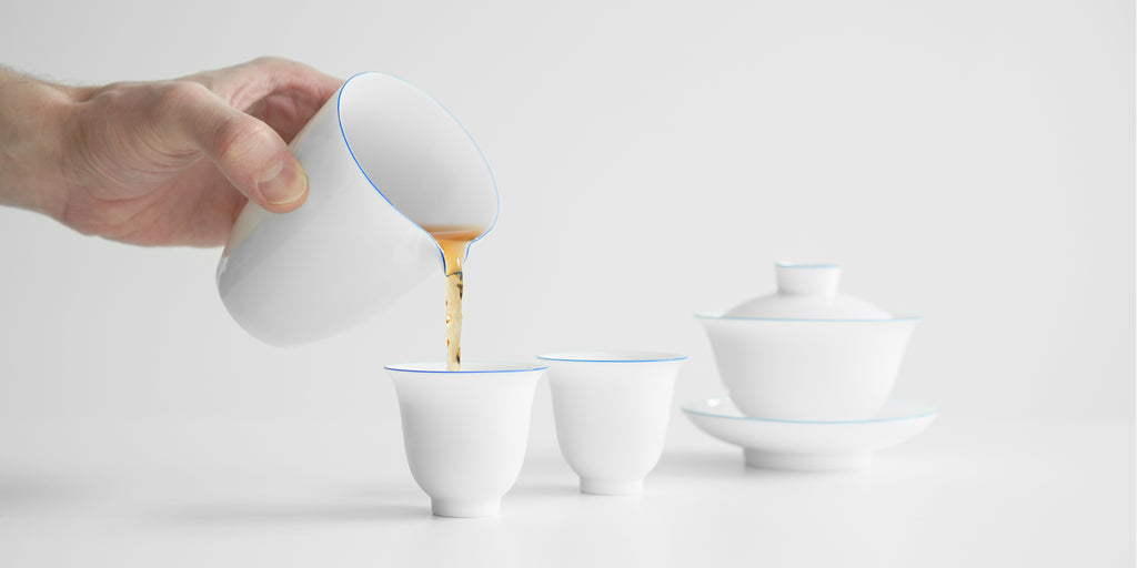 A hand pours tea from a white porcelain server into a white teacup. To the right there is a gaiwan, also white with blue rings painted around the edges.