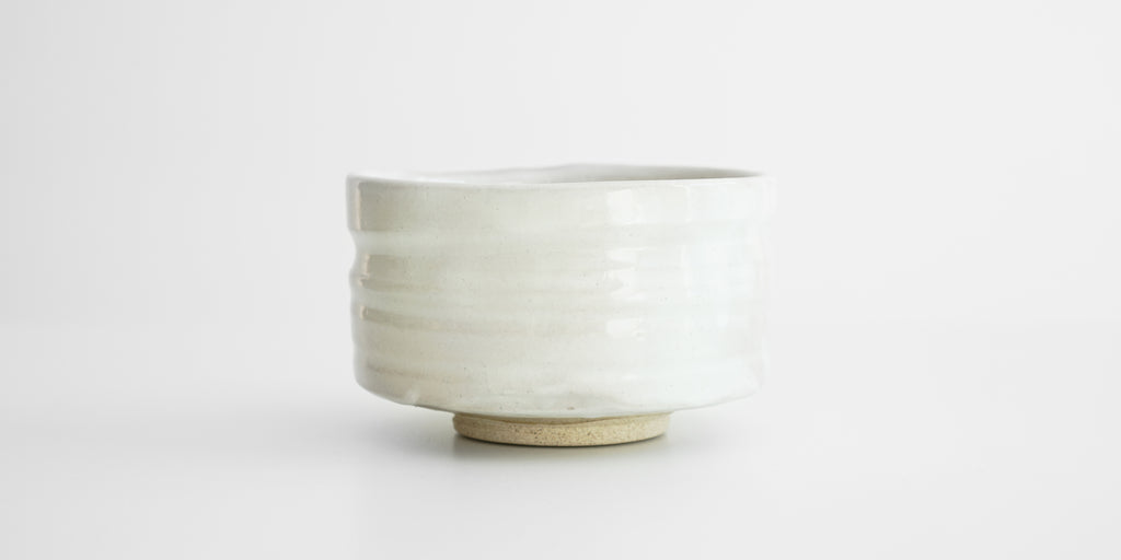 Side view of a straight-sided white teabowl. The foot is unglazed, and the sides have some indentations going around them for the hands to fit into.