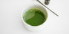 A white bowl with matcha brewed placed on a white surface. A matcha scoop and whisk are in the background.