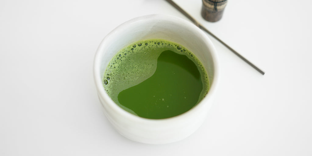 A white bowl with matcha brewed placed on a white surface. A matcha scoop and whisk are in the background.