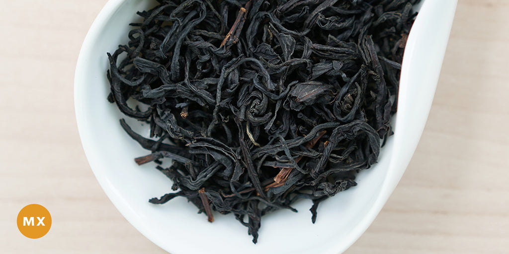A white dish containing dry black tea leaves. An orange circle in the bottom left corner reads "MX"