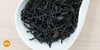 A white dish containing dry black tea leaves. An orange circle in the bottom left corner reads "MX"