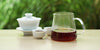 From left to right, a gaiwan, two teacups, and a glass server. The teacups and server have black tea in them.