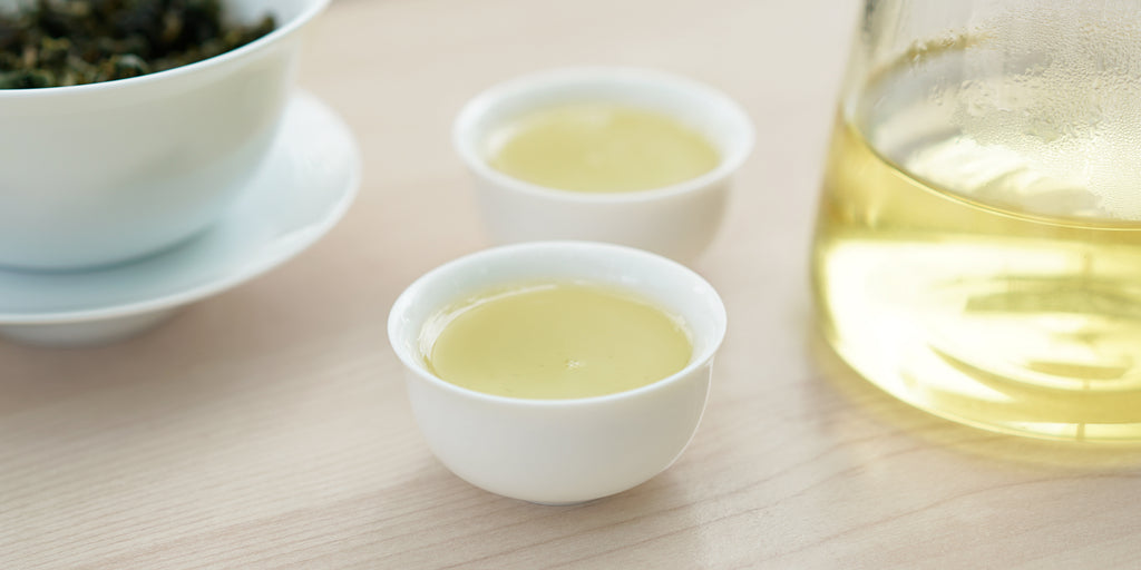 Two small teacups of Formosa oolong tea, with a gaiwan in the background on the left and a glass server on the right.