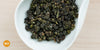 A white dish containing dry Formosa oolong tea leaves. An orange circle in the bottom left corner reads "MX"