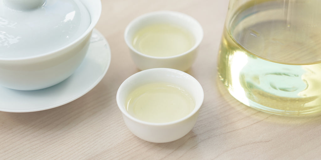 Two small teacups of green tea, with a gaiwan in the background on the left and a glass server on the right.