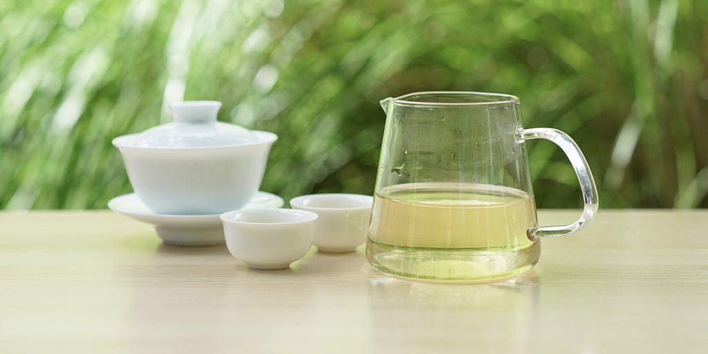 From left to right, a gaiwan, two teacups, and a glass server. The teacups and server have green tea in them.