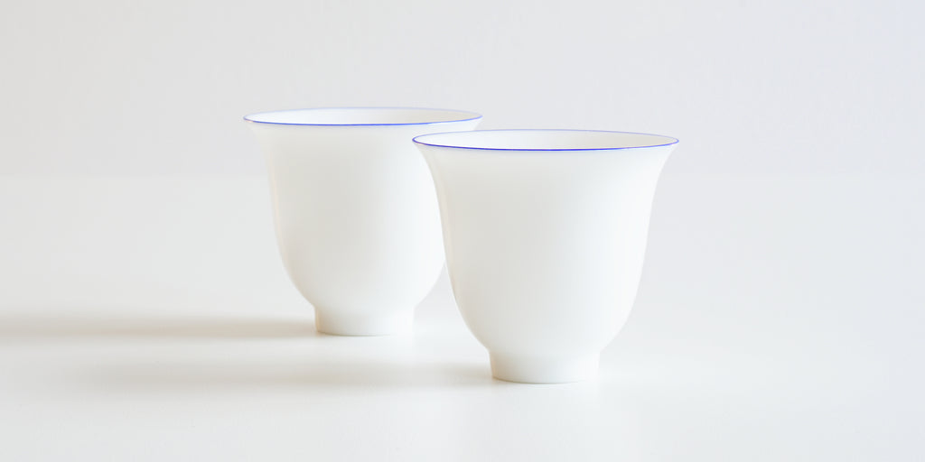 Two white teacups, curving up at the base and out towards the thin lip, with a blue ring painted around the edge.