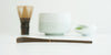 Close up of a purple bamboo matcha scoop, with a whisk, white bowl, and dish full of matcha out of focus in the background.