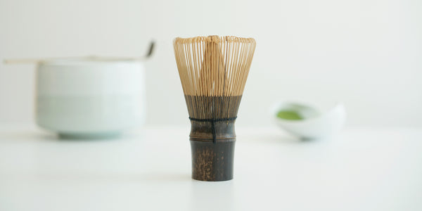 A purple bamboo matcha whisk on a white surface, with a white bowl, matcha scoop, and white dish full of matcha out of focus in the background.