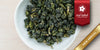 A white dish containing dry Formosa oolong tea leaves. A red ribbon in the top right corner reads “Red Label Collection”, and a gold ribbon in the bottom right corner reads “New Arrival”