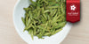 A white dish containing dry green tea leaves. A red ribbon in the top right corner reads “Red Label Collection”