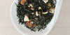 A white dish containing dry green tea leaves, dried orange peel, and ginger.