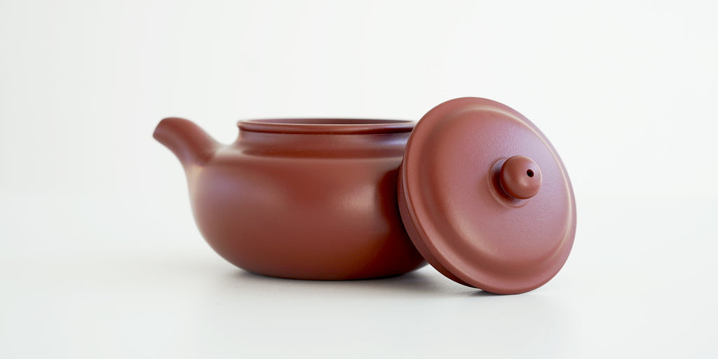 Side view of a terracotta-colored yixing teapot. The lid is facing the camera, resting against the handle.