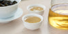 Two small teacups of Tieguanyin tea, with a gaiwan in the background on the left and a glass server on the right.