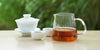 From left to right, a gaiwan, two teacups, and a glass server. The teacups and server have black tea in them.