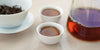 Two small teacups of black tea, with a gaiwan in the background on the left and a glass server on the right.