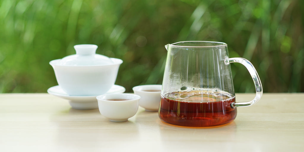 From left to right, a gaiwan, two teacups, and a glass server. The teacups and server have black tea in them.