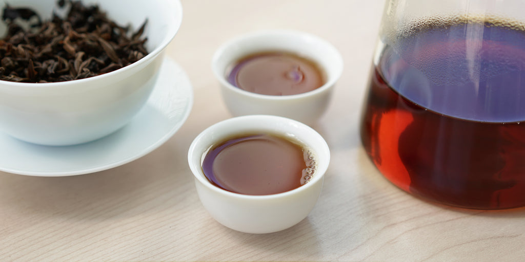 Two small teacups of black tea, with a gaiwan in the background on the left and a glass server on the right.