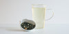 A white dish of dry green tea leaves, dried orange peel, and ginger. A glass cup of brewed tea is in the background.