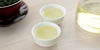 Two small teacups of Formosa oolong tea, with a gaiwan in the background on the left and a glass server on the right.