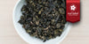 A white dish containing dry Tieguanyin tea leaves. A red ribbon in the top right corner reads “Red Label Collection”