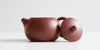Side view of a terracotta-colored yixing teapot. The lid is facing the camera, resting against the handle.