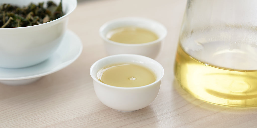 Two small teacups of Formosa oolong tea, with a gaiwan in the background on the left and a glass server on the right.