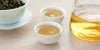Two small teacups of Formosa oolong tea, with a gaiwan in the background on the left and a glass server on the right.