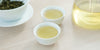 Two small teacups of green tea, with a gaiwan in the background on the left and a glass server on the right.