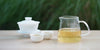 From left to right, a gaiwan, two teacups, and a glass server. The teacups and server have green tea in them.