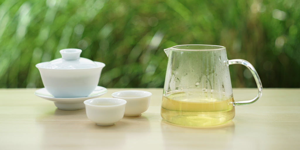 From left to right, a gaiwan, two teacups, and a glass server. The teacups and server have green tea in them.