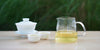 From left to right, a gaiwan, two teacups, and a glass server. The teacups and server have green tea in them.