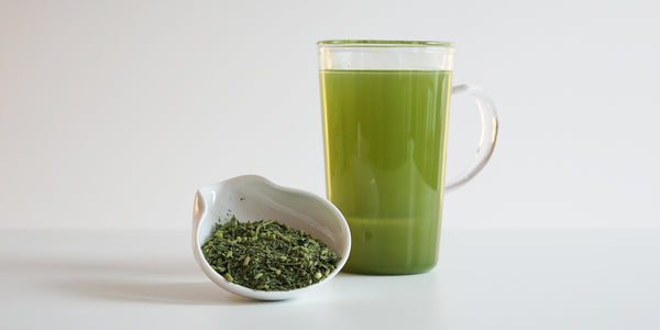 A white tea dish with dry green tea leaves, toasted rice and matcha powder. A glass cup full of brewed genmai matcha is in the background.