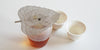 Metal leaf strainer in the shape of a bodhi leaf over a glass server of tea. Two white teacups in the background.