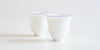 Side view of a pair of white porcelain teacups, with a ring of blue painted around the rim.
