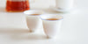 Side view of a pair of white teacups with a blue ring around the rim, each filled with tea. A glass server filled with tea and a gaiwan are out of focus in the background.
