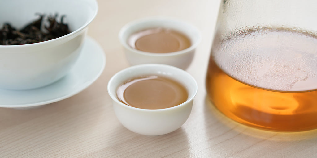 Two small teacups of Phoenix oolong tea, with a gaiwan in the background on the left and a glass server on the right.