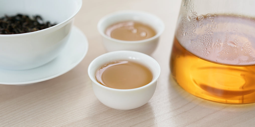 Two small teacups of Phoenix oolong tea, with a gaiwan in the background on the left and a glass server on the right.