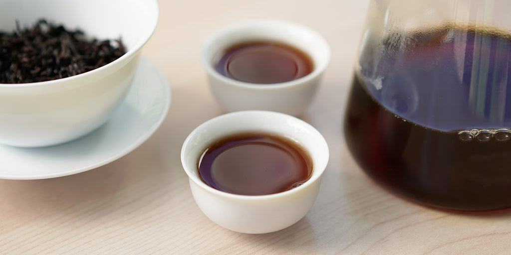 Two small teacups of pu-erh tea, with a gaiwan in the background on the left and a glass server on the right.