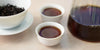 Two small teacups of pu-erh tea, with a gaiwan in the background on the left and a glass server on the right.