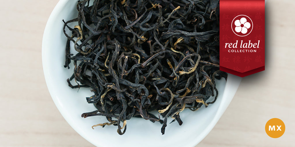 A white dish containing dry black tea leaves. A red ribbon in the top right corner reads “Red Label Collection”, and an orange circle in the bottom right corner reads “MX”
