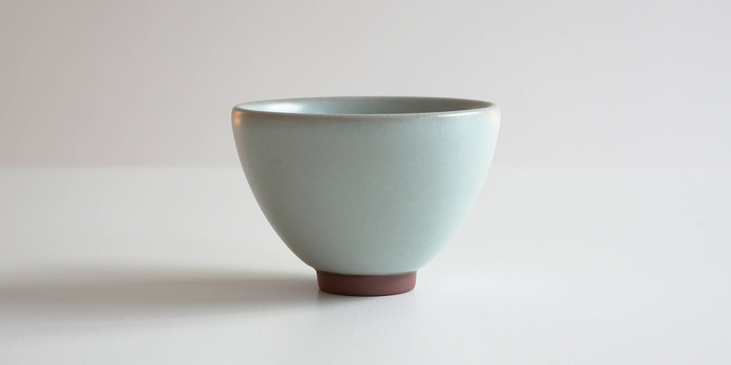 Side view of a blue-green celadon teacup, showing the oval shape and unglazed foot.