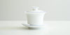 Side view of a white porcelain gaiwan, with blue rings painted around the edges of the plate, bowl, and the handle of the lid. The sides start straight before curving out towards the lip.