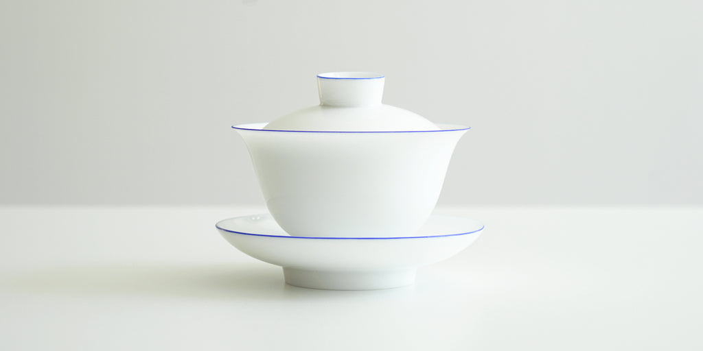 Side view of a white porcelain gaiwan, with blue rings painted around the edges of the plate, bowl, and the handle of the lid. The sides are rounded at the bottom before curving out towards the lip.